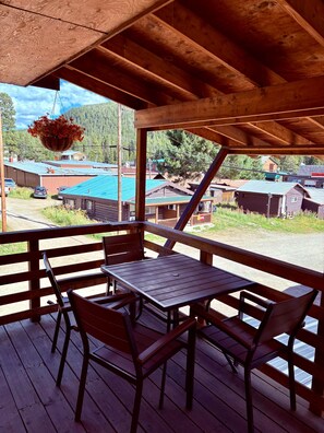 A wooden deck with a table and chairs, overlooking a rural area with houses and a hanging flower basket. - Miller Mountain Cabin (Cooke City)