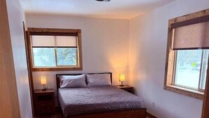 Une chambre avec un plancher en bois, un lit avec une tête de lit, deux tables de chevet avec des lampes et une fenêtre donnant sur un paysage enneigé.