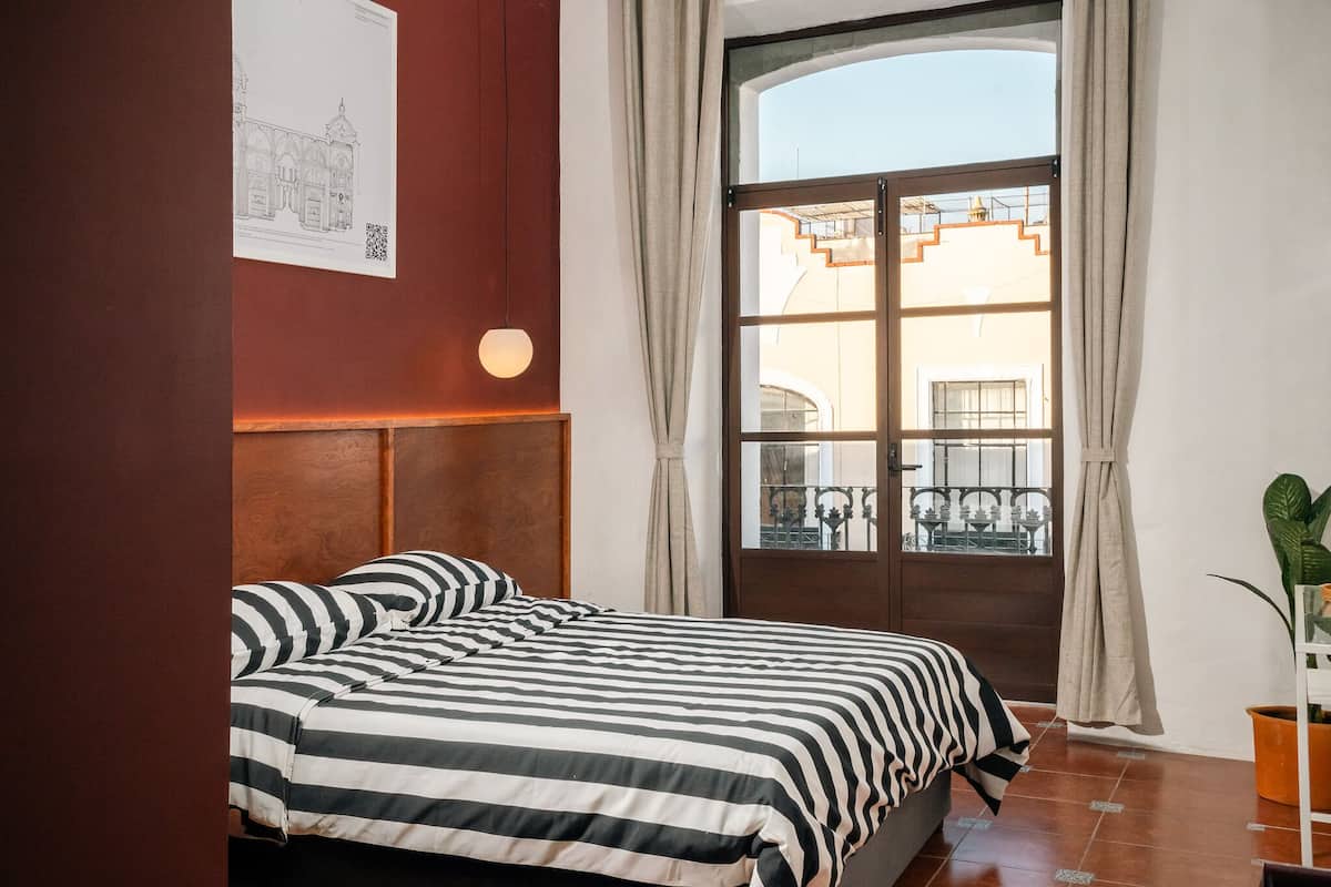 Une chambre avec une grande fenêtre, un lit avec une literie à rayures et un luminaire fixé au mur.