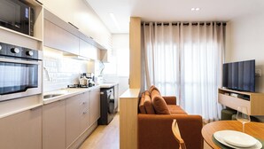 Une cuisine moderne dotée d’appareils électroménagers intégrés, un coin repas avec une table dressée pour deux personnes, et un salon avec un canapé et un téléviseur.