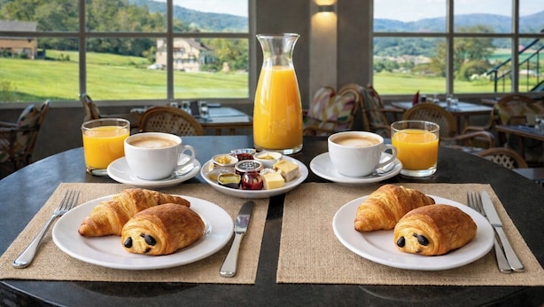 A breakfast setting with croissants, coffee, juice, and a view of a green landscape through large windows.
