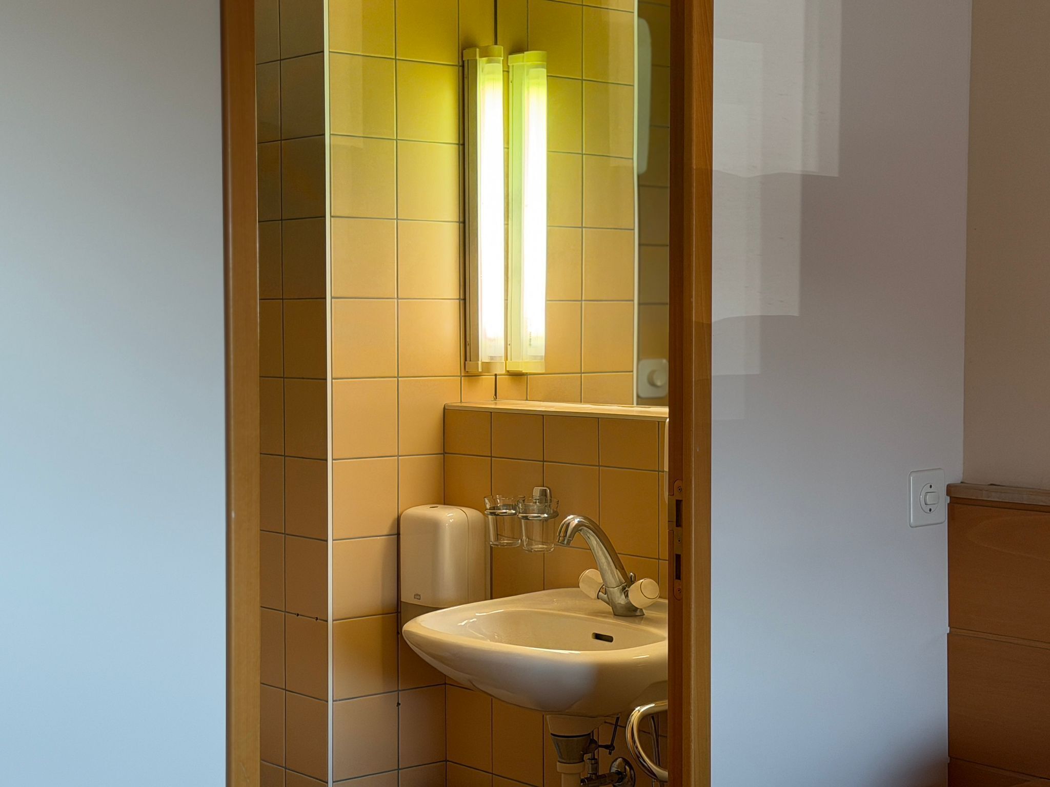 Ein Badezimmer mit einem weißen Waschbecken, einem Spiegel und einer gelb gefliesten Wand.