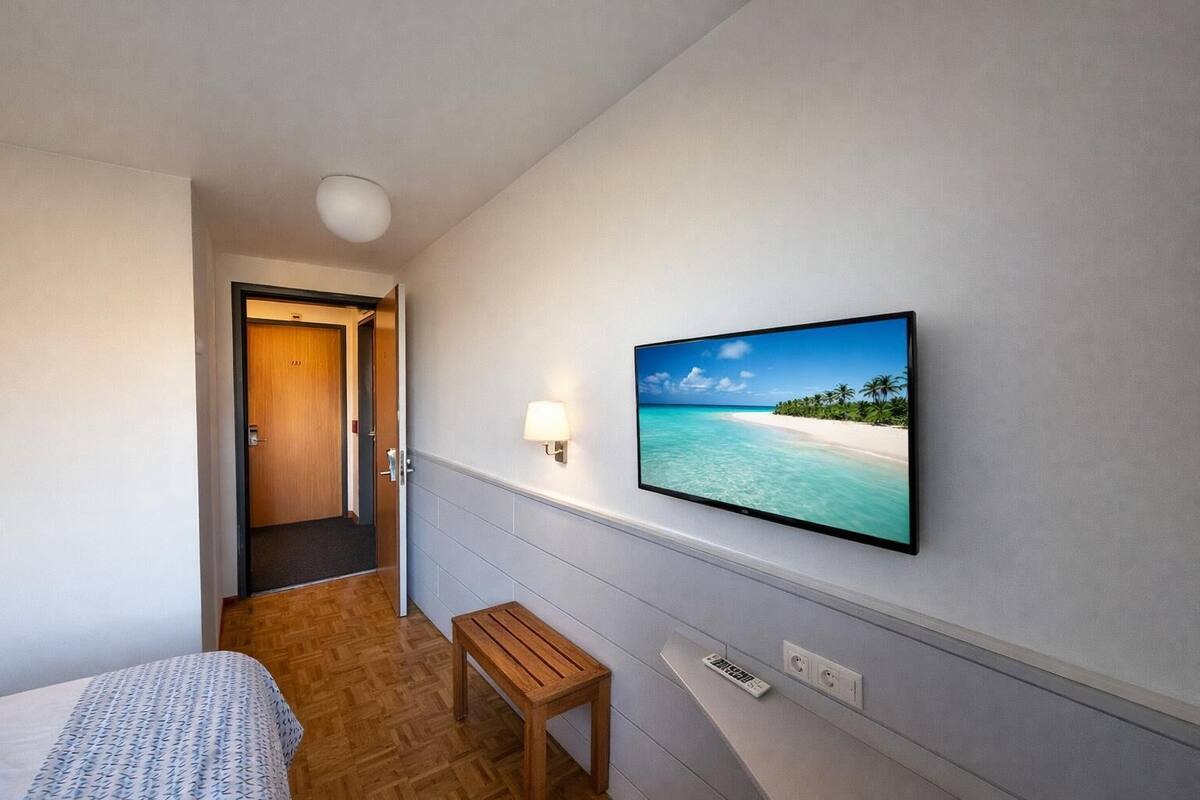 Une chambre d’hôtel avec un lit, un banc en bois, un téléviseur fixé au mur qui diffuse une scène de plage, et une porte donnant sur une autre pièce.