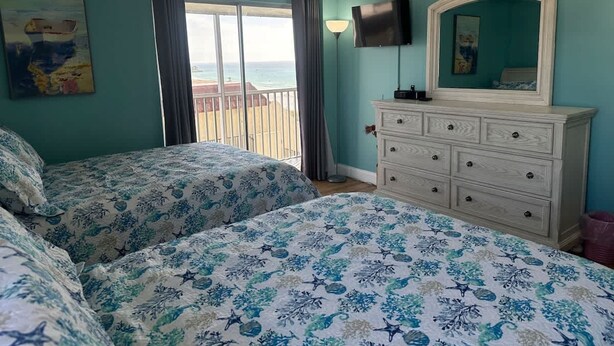 Een slaapkamer met twee bedden, een dressoir, een plafondventilator, een televisie en een balkon met uitzicht op de oceaan.