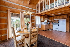 A wooden cabin with a dining area featuring a round table and chairs, a kitchen with wooden cabinets and appliances, and a balcony with a railing. - Super View Smoky Mtn Log Cabin Pool Table Hot Tub (Sevierville)