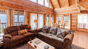 A cozy wooden cabin with a large living area featuring two sofas, a coffee table, and a dining table. Large windows offer scenic mountain views. - Super View Smoky Mtn Log Cabin Pool Table Hot Tub (Sevierville)