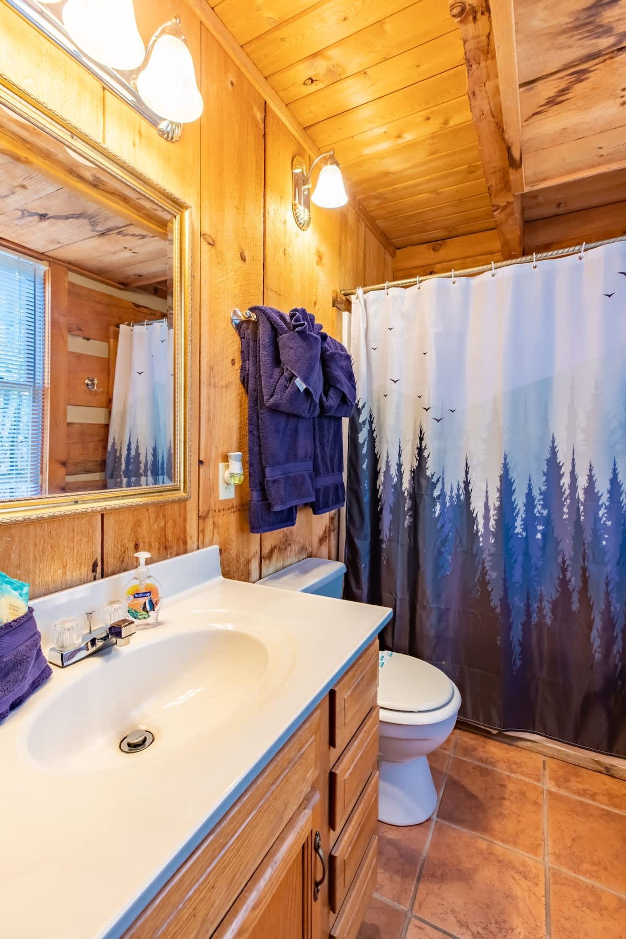 A wooden cabin bathroom with a large mirror, a sink, a toilet, and a shower curtain with a forest design.