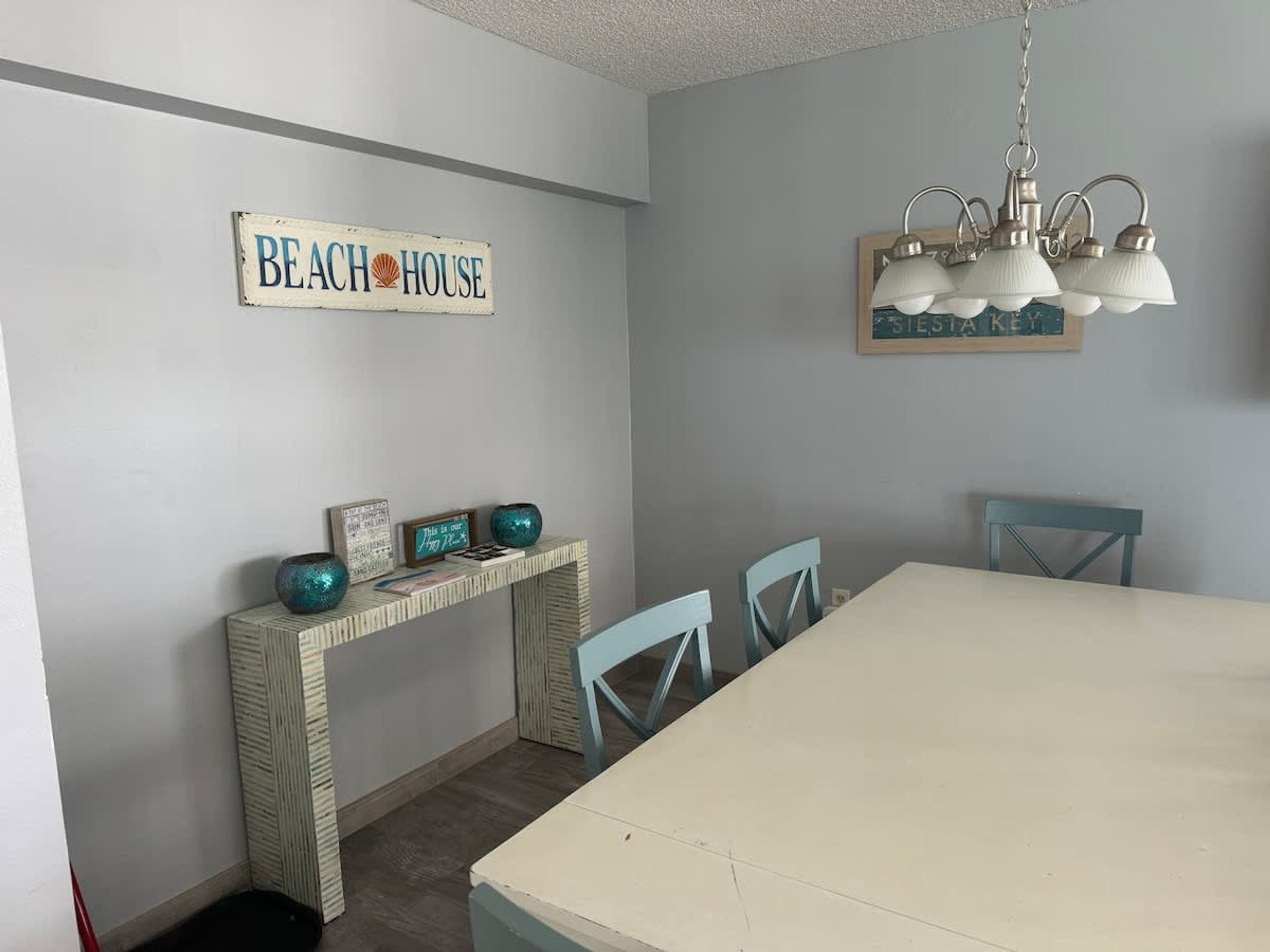 A dining area with a large table, chairs, a console table with decorative items, a sign reading "BEACH HOUSE", and a hanging light fixture.