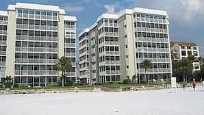 A modern building with multiple floors and large windows, surrounded by a sandy area with some greenery. - Beach Pool Open Direct Beachfront Condo Sunsets (Siesta Key)