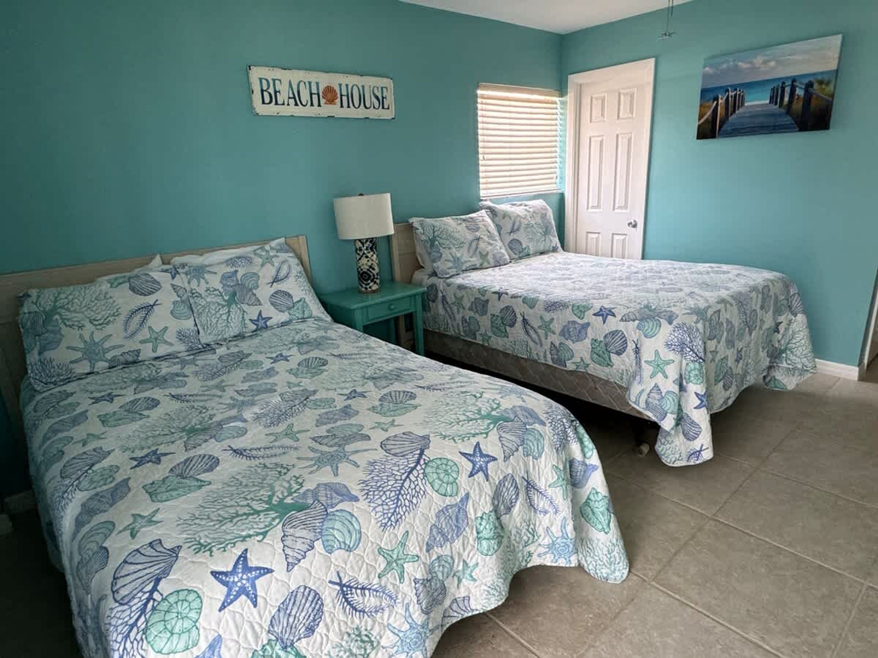 Un dormitorio con dos camas, cada una con una colcha náutica azul y blanca. Hay un letrero en la pared que dice 'BEACH HOUSE' y un cuadro con una escena de playa.