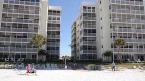 Hotel al costat de la platja, amb diverses plantes, balcons i una zona de platja de sorra amb gandules.