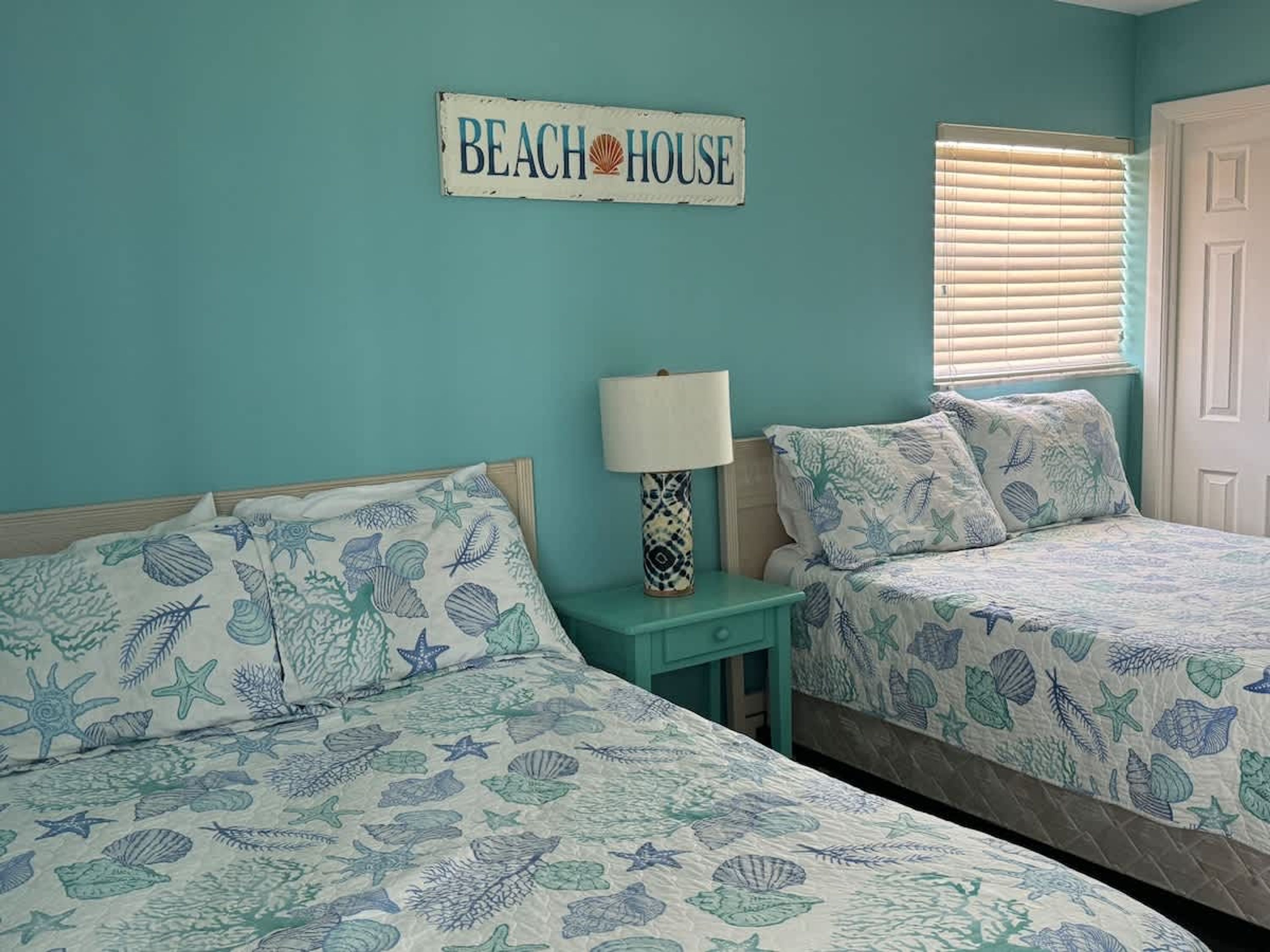 Une chambre avec deux lits, chacun recouvert d’une couvre-lit sur le thème de la mer. Une pancarte sur le mur indique : « MAISON DE PLAGE ».