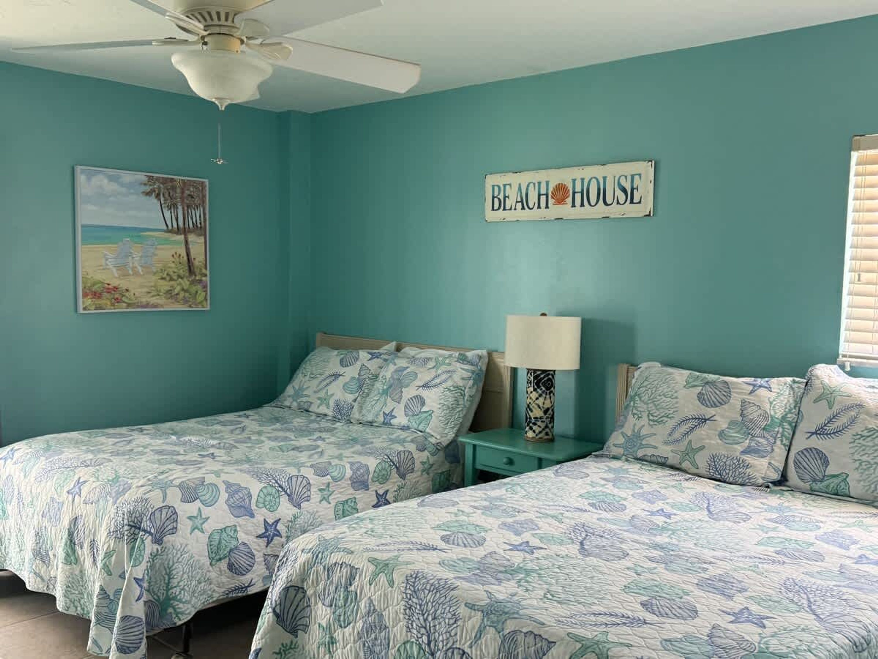 Een slaapkamer met twee bedden, een plafondventilator, een schilderij met een strandthema, en een bordje met de tekst 'BEACH HOUSE'.