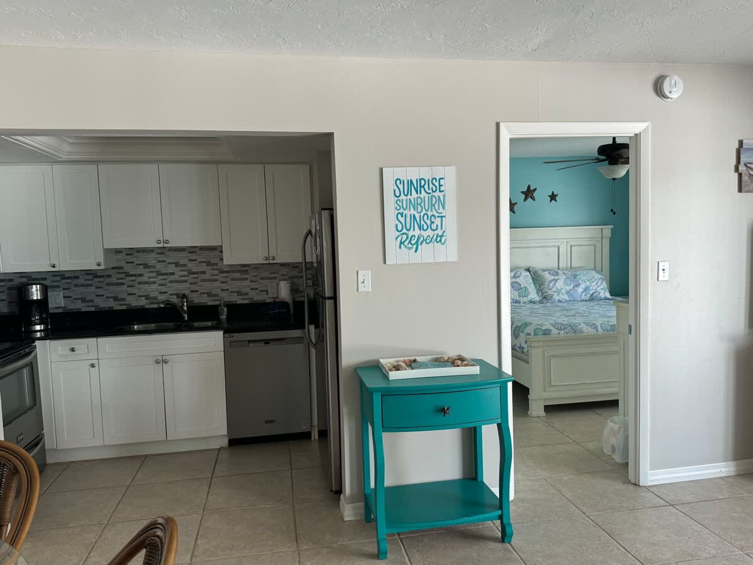 Une cuisine compacte avec des meubles blancs, un plan de travail noir et une petite table d’appoint turquoise agrémentée d’un objet décoratif. Un panneau mural porte l’inscription : « LEVER DE SOLEIL, COUP DE SOLEIL, COUCHER DE SOLEIL. Répéter. »