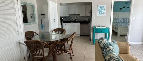 A dining area with a glass table and wicker chairs, a kitchen with white cabinets, and a teal side table.