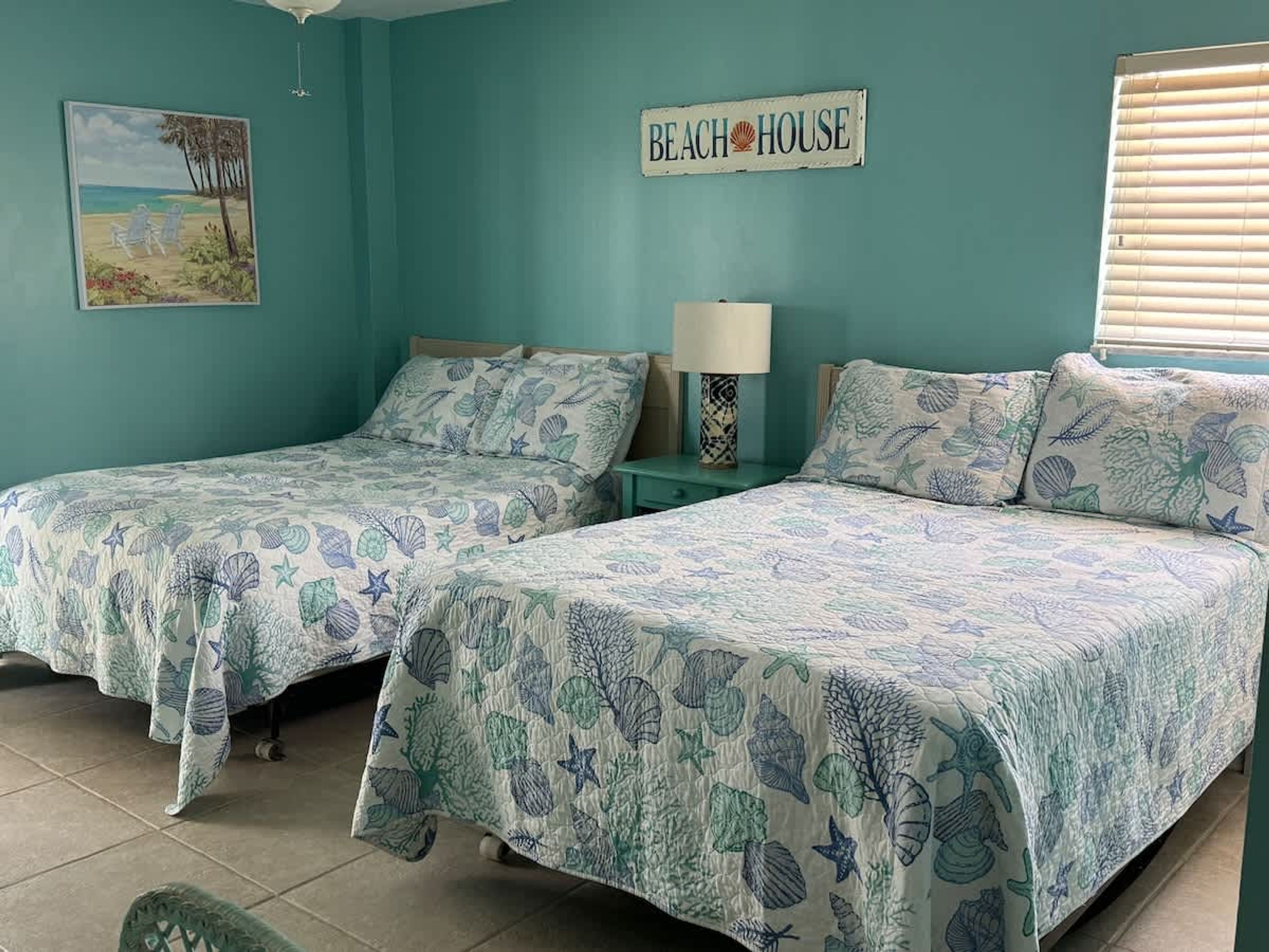 Une chambre avec deux lits, chacun recouvert d’une couvre-lit sur le thème de la plage, une table de chevet avec une lampe, une pancarte indiquant « MAISON DE PLAGE » et un tableau représentant une scène de plage accroché au mur.