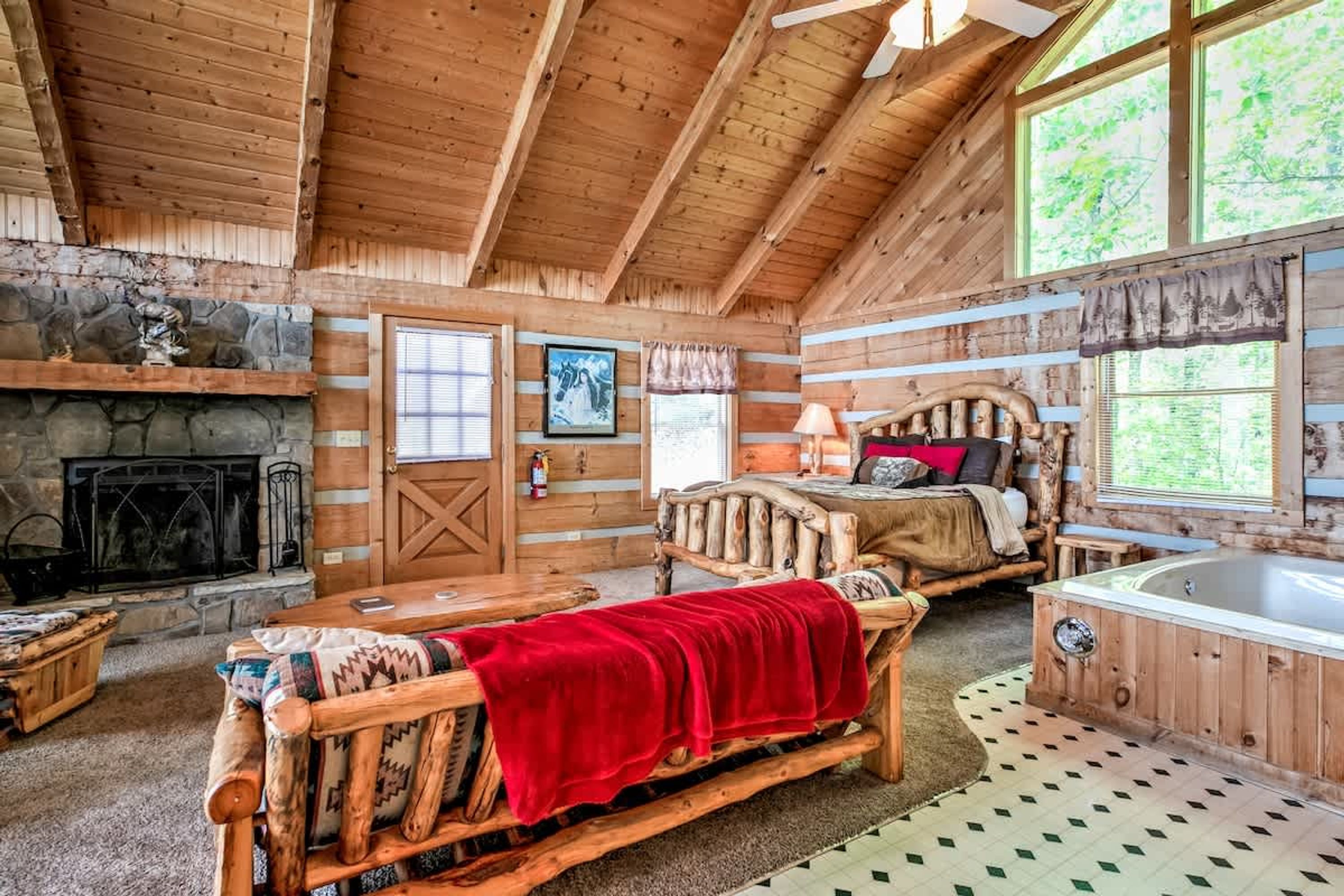 A log cabin interior with a stone fireplace, a bed with a red blanket, a wooden bench, and a hot tub.