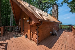 A wooden cabin with a sloped roof and a stone chimney, featuring a deck with outdoor seating and a view of the surrounding forest. - Cute Studio Cabin Views Private Hot Tub Relax (Sevierville)