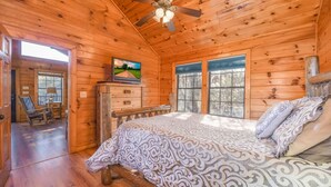 A wooden cabin bedroom with a bed, a ceiling fan, a dresser, a television, and a window. - Secluded Cabin Near Smoky Mountains Hot Tub Honeymoon (Sevierville)