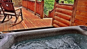 A wooden deck with a hot tub, a rocking chair, and a view of lush greenery. - Secluded Cabin Near Smoky Mountains Hot Tub Honeymoon (Sevierville)