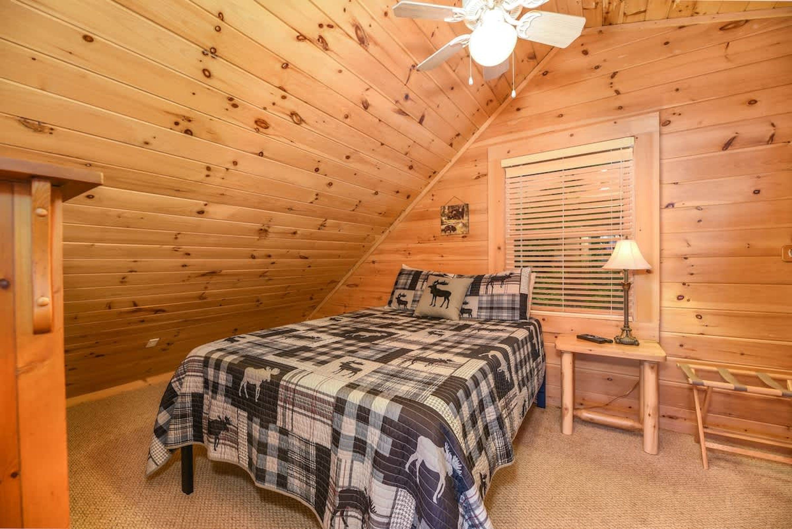 Un dormitorio acogedor con techo de madera, una cama con manta a cuadros, una mesita de noche con lámpara y una ventana con persianas.