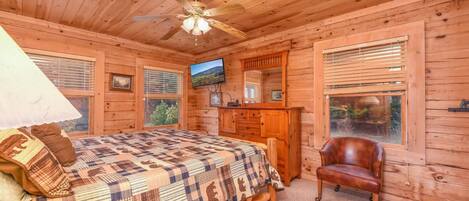 A wooden cabin bedroom with a bed, a desk, a chair, and a ceiling fan.