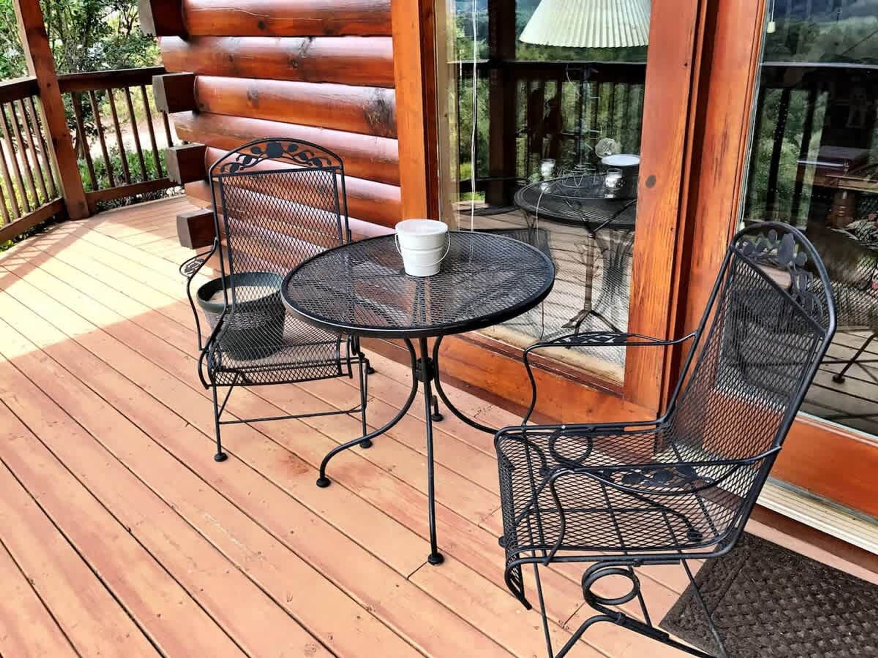 Une terrasse en bois avec une table ronde en métal et deux chaises, une tasse blanche posée sur la table, et une porte vitrée donnant sur un espace patio.
