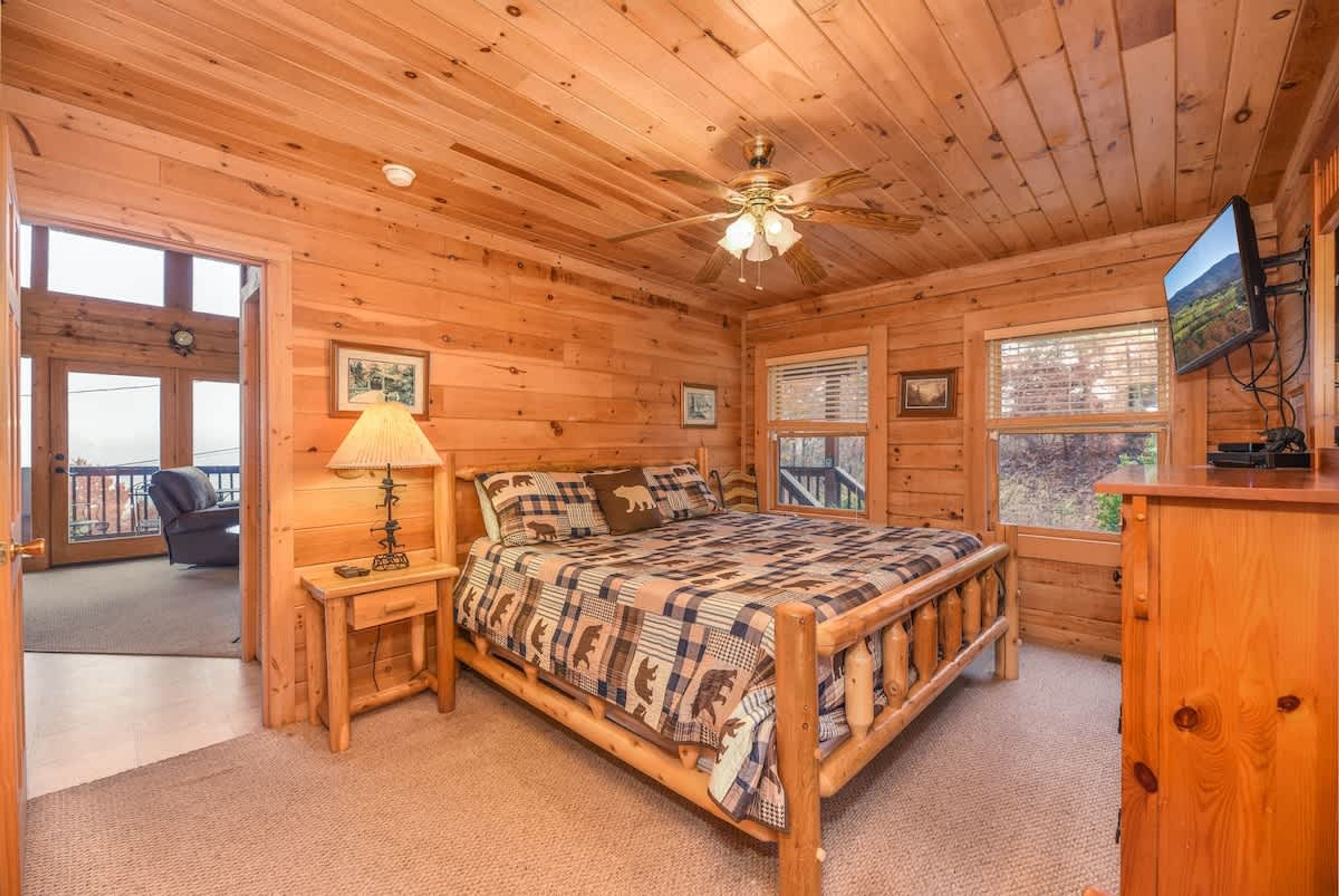 A wooden cabin bedroom with a bed, a nightstand, a lamp, a dresser, and a TV.