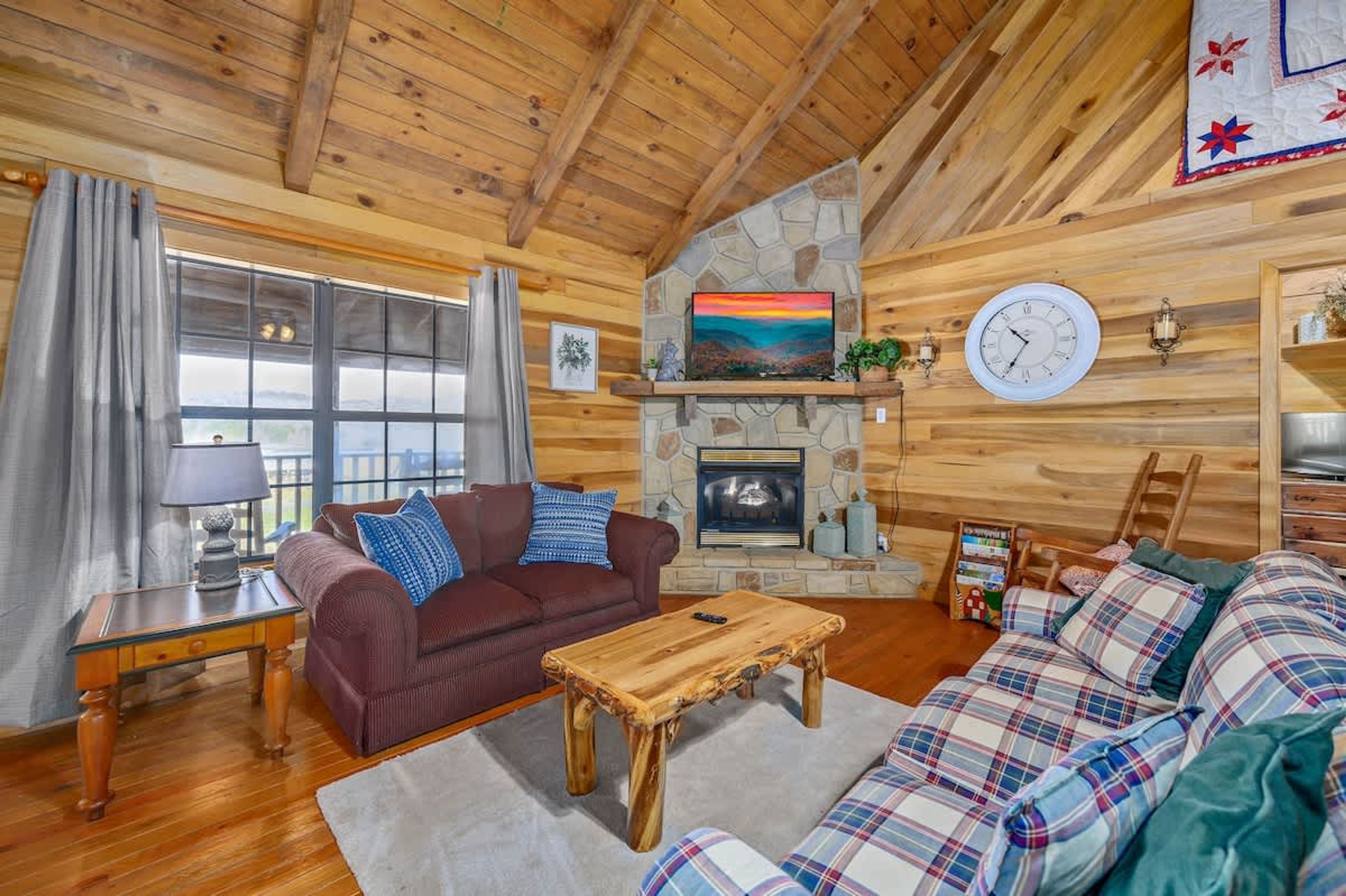 A cozy cabin interior with a stone fireplace, wooden walls, a large window, and a plaid sofa.