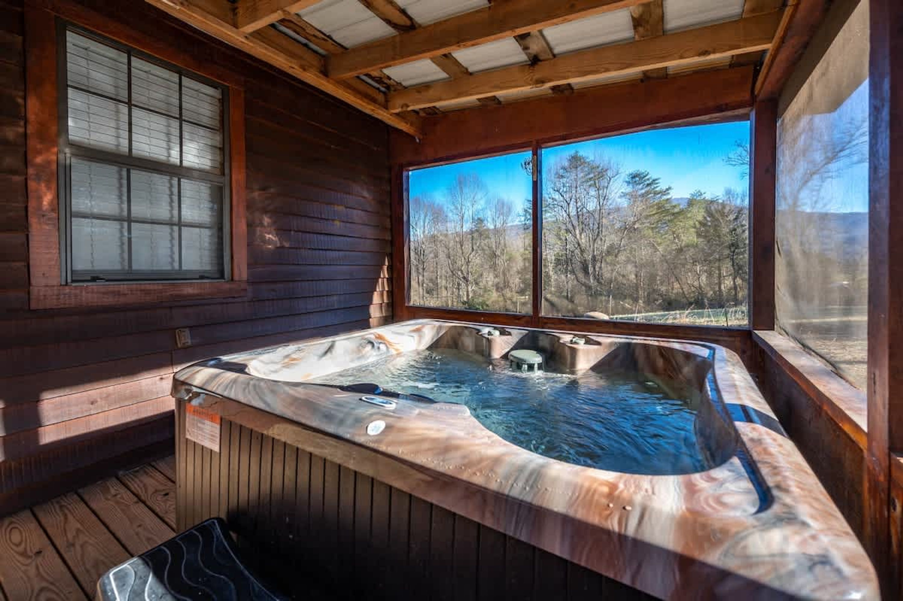A wooden cabin with a hot tub and large windows overlooking a forest.