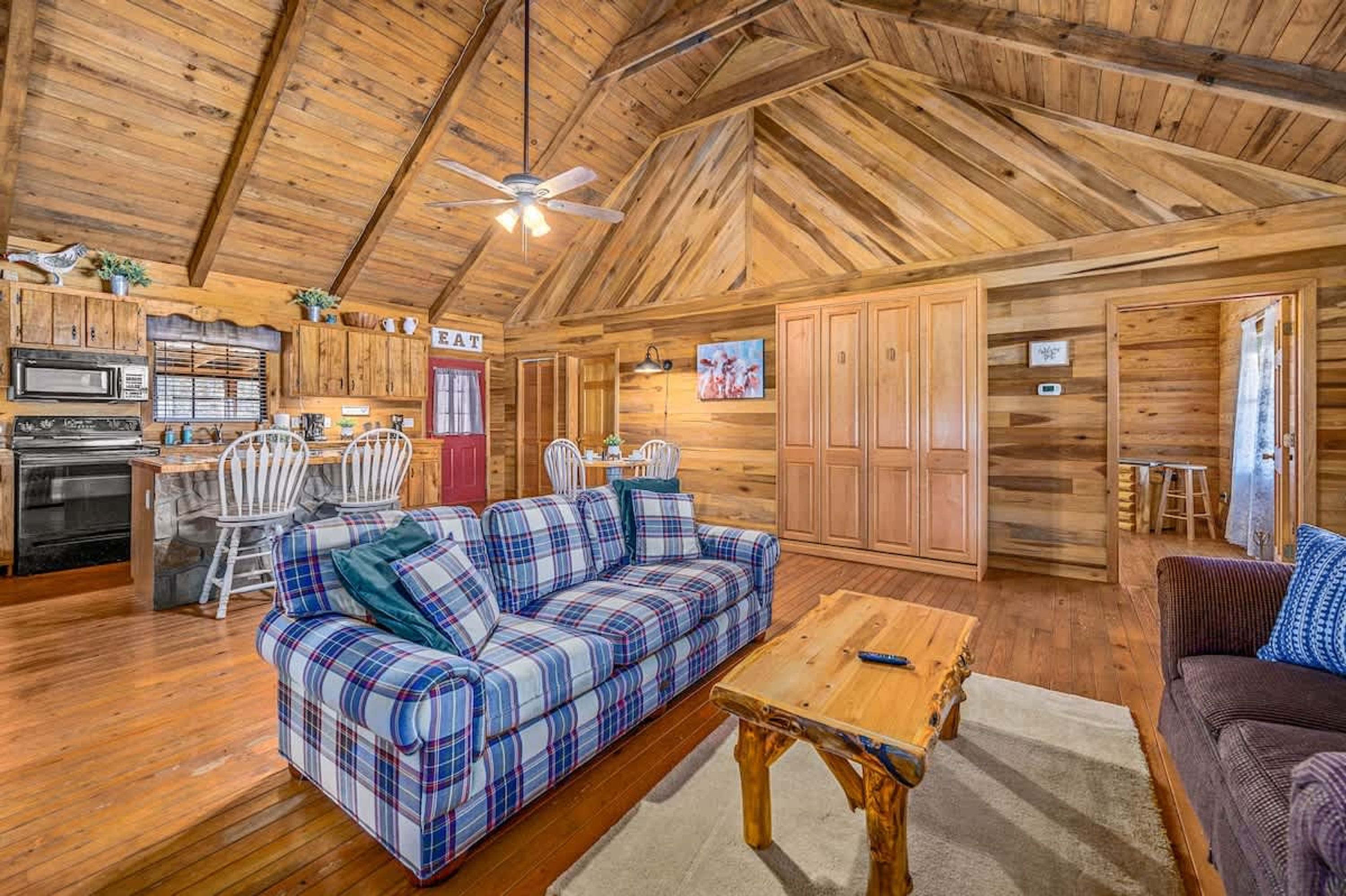 A log cabin interior with a plaid sofa, wooden dining table, and a kitchen area with a microwave and chairs.