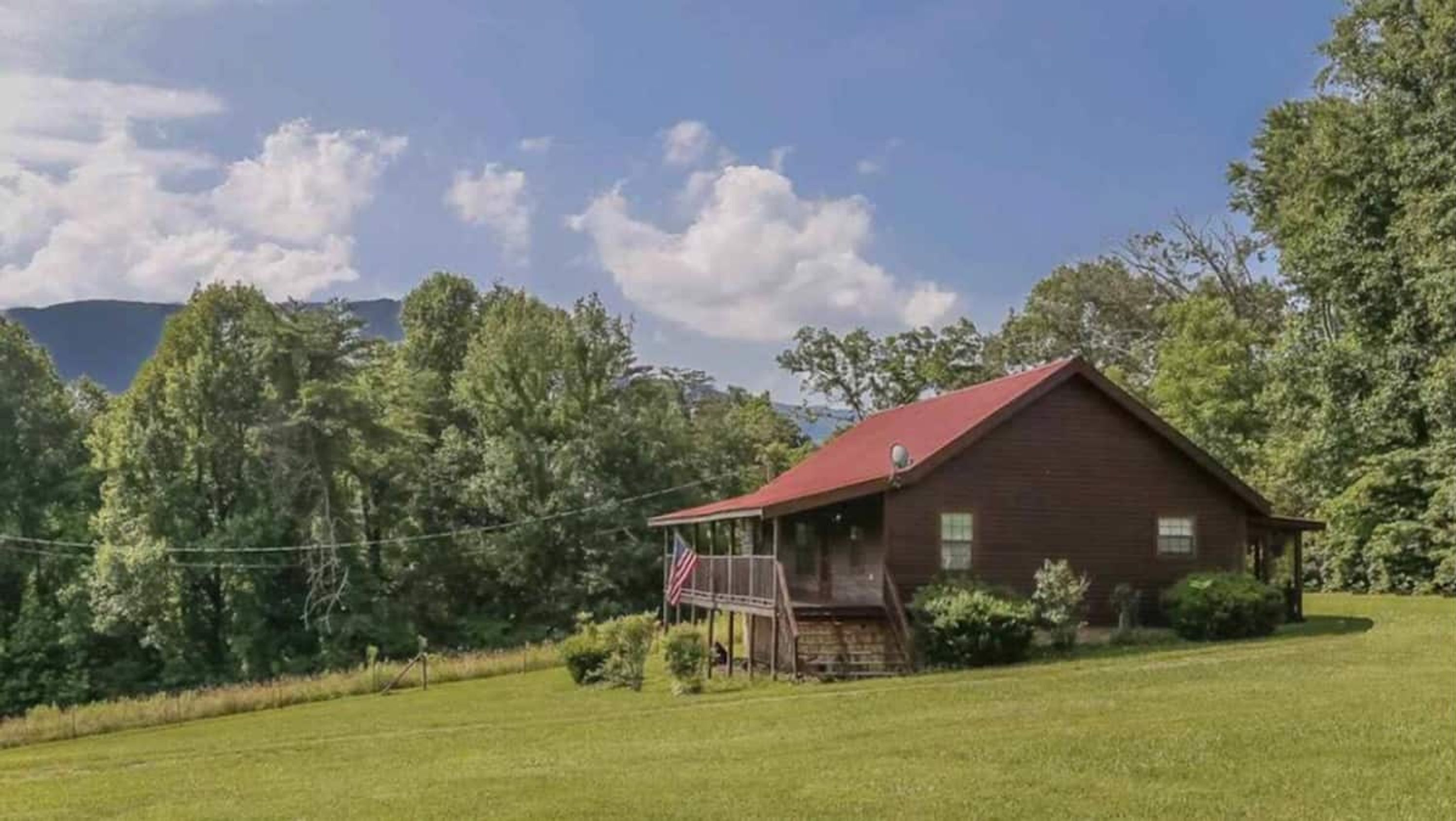 A wooden cabin with a red roof, surrounded by a grassy field and trees, with mountains in the background.