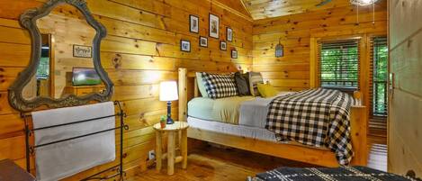 Dormitorio de cabaña de madera con cama, espejo, cuadros enmarcados, ventilador de techo y una mesita de noche con lámpara.