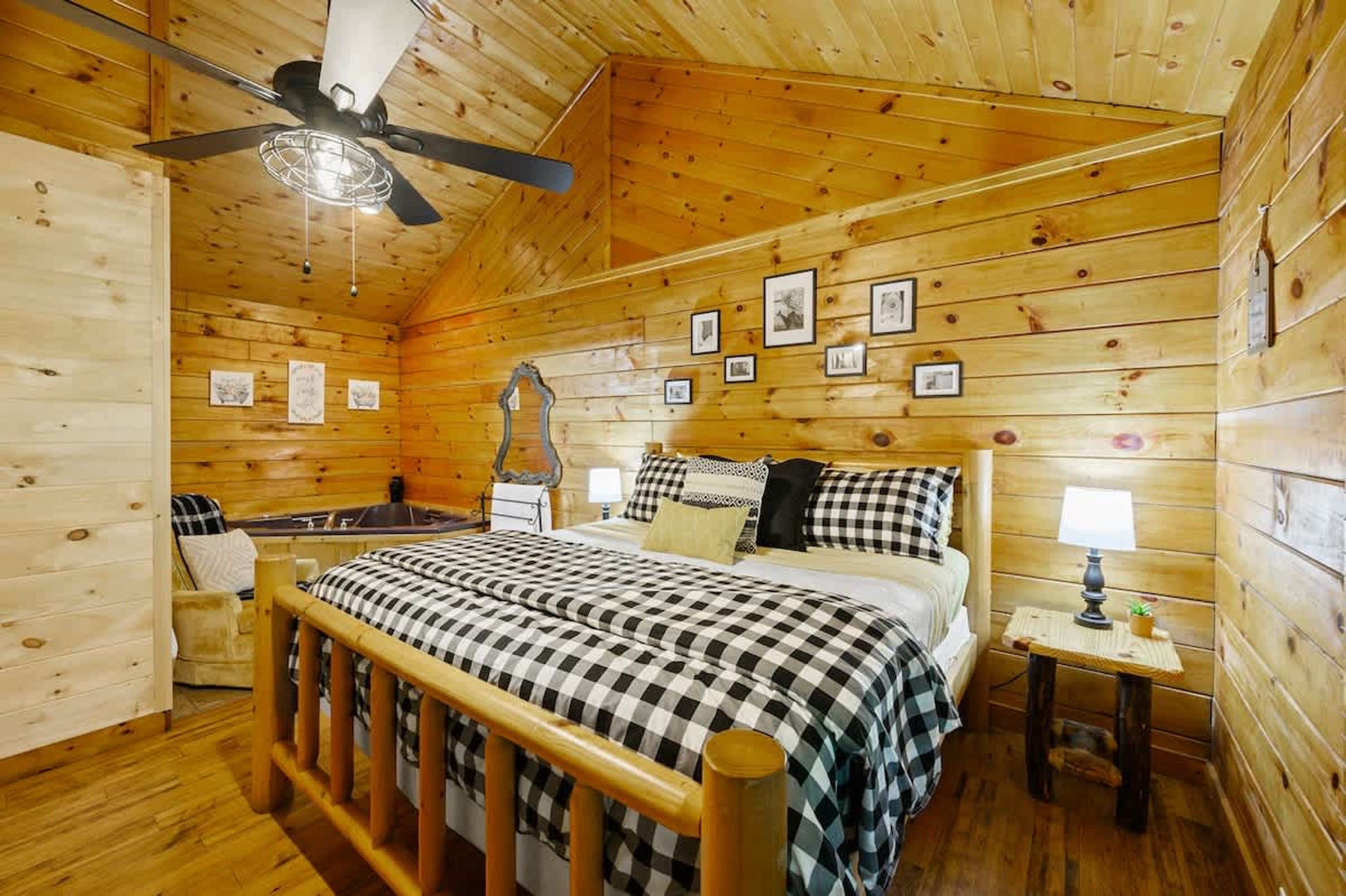 Een slaapkamer in een houten hut met een bed, een plafondventilator, ingelijste foto’s aan de muur en een tafeltje met een lamp.