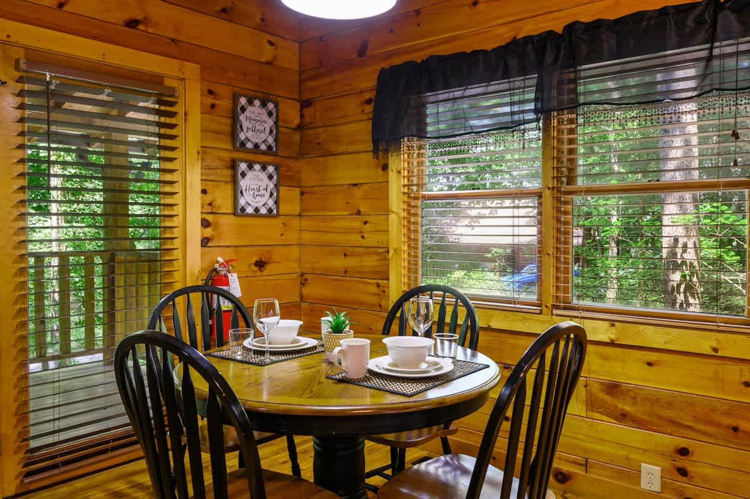 Interior de uma cabana de madeira com uma mesa de jantar posta para quatro pessoas, rodeada por cadeiras. Há duas janelas com persianas, uma delas com uma cortina decorativa.