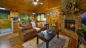 A cozy cabin interior with a stone fireplace, wooden walls, a leather sofa, and a television. - Location Log Cabin Hot Tub Fireplace Pool Fishing (Sevierville)