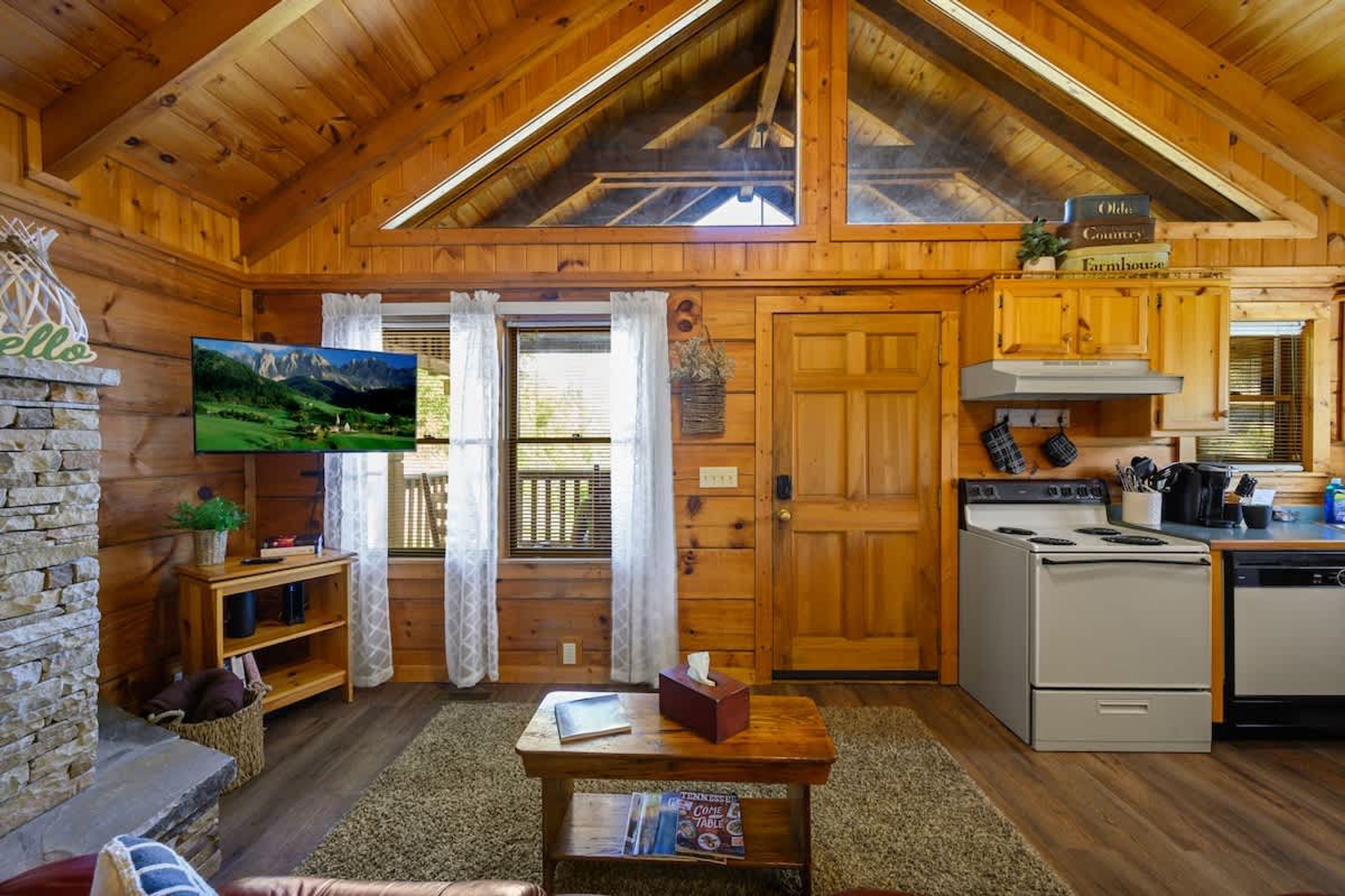 A cozy cabin interior with wooden walls, a stone fireplace, a television, a kitchen with a stove and oven, and a dining area with a table and chairs.