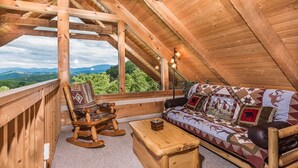 A cozy wooden loft bedroom with a large window, a rocking chair, a sofa with patterned cushions, and a small wooden chest. - View Private Hot Tub Pool Table Fireplace Relax (Sevierville)