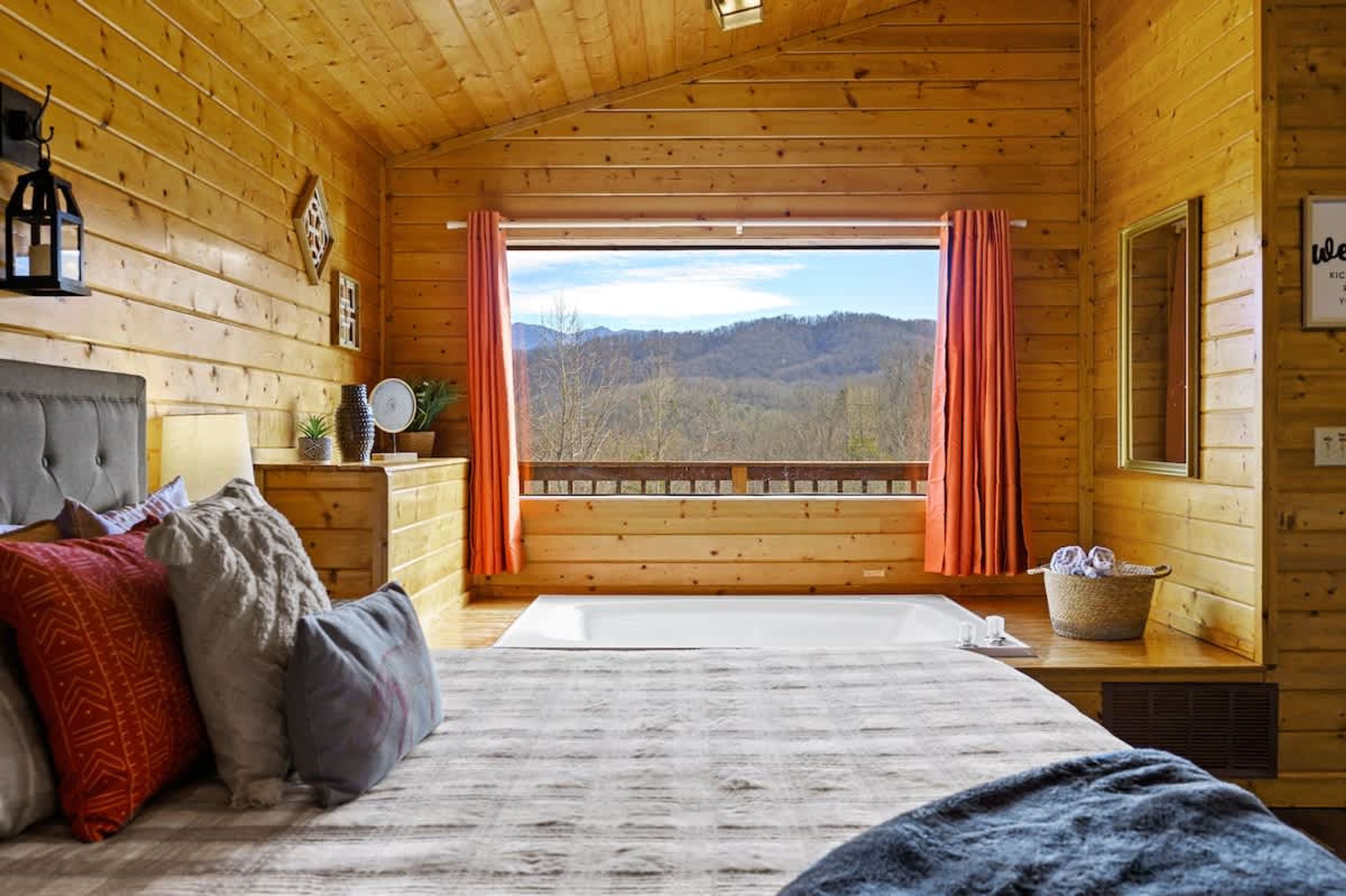 A wooden cabin bedroom with a bed, pillows, a nightstand, a window with curtains, and a view of a forested landscape.