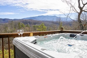 Ein Whirlpool mit zwei Weingläsern auf einer Holzdiele mit Blick auf eine gebirgige Landschaft.