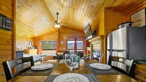 A wooden cabin interior with a dining table set for two, a ceiling fan, a kitchen area with a refrigerator, and a large window. - Great View Location Studio Log Cabin Hot Tub (Gatlinburg)