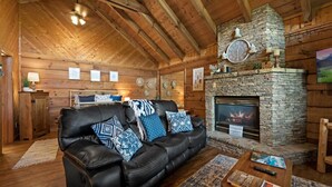 A cozy log cabin interior with a stone fireplace, leather sofa, and wooden beams.