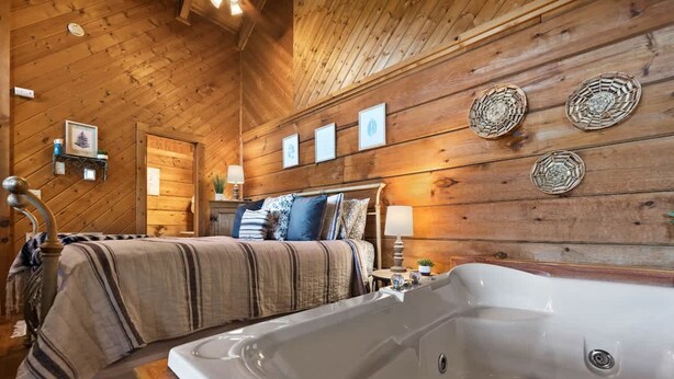 A cozy wooden cabin bedroom with a large bathtub, a bed with striped bedding, and wall-mounted decor.