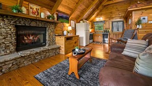 A cozy cabin interior with a stone fireplace, wooden ceiling beams, a kitchen area, and a living room with a brown leather sofa. - Private Studio Log Cabin Hot Tub Fireplace Relax (Sevierville)