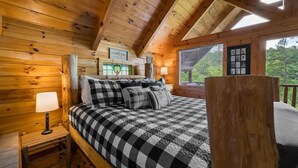 A wooden cabin bedroom with a checkered bedspread, a nightstand with a lamp, and a window with blinds. - Private Studio Log Cabin Hot Tub Fireplace Chill (Sevierville)
