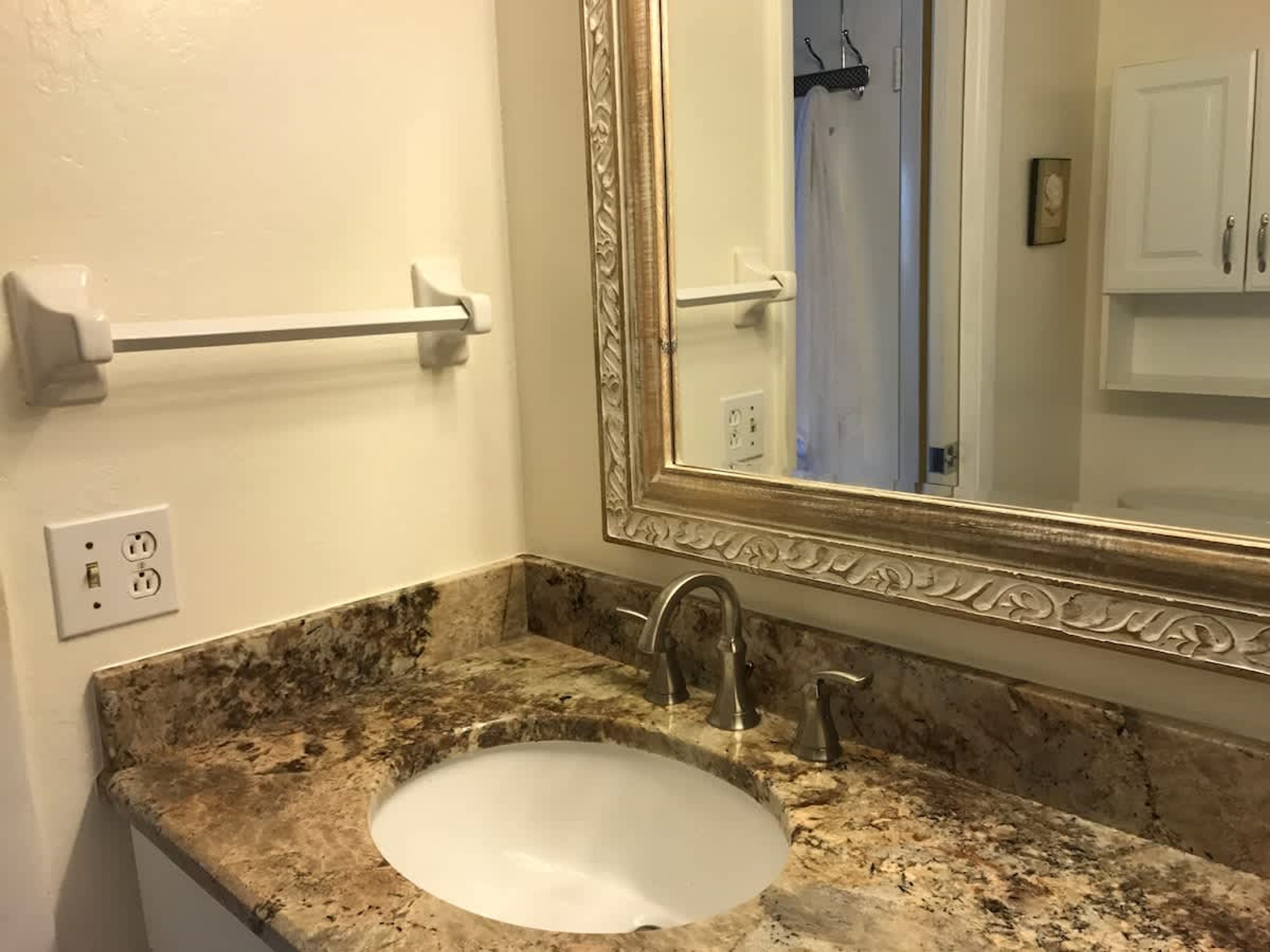 A bathroom with a granite countertop sink, a large mirror with an ornate frame, a towel rack, and a towel dispenser.