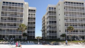 Un hotel frente a la playa con varios pisos, balcones y una zona de arena con sillas de descanso.