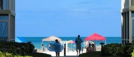 Ein Weg führt zu einem Strandabschnitt, wo eine Person ein Surfbrett trägt, Zelte stehen und an beiden Seiten Gebäude zu sehen sind.