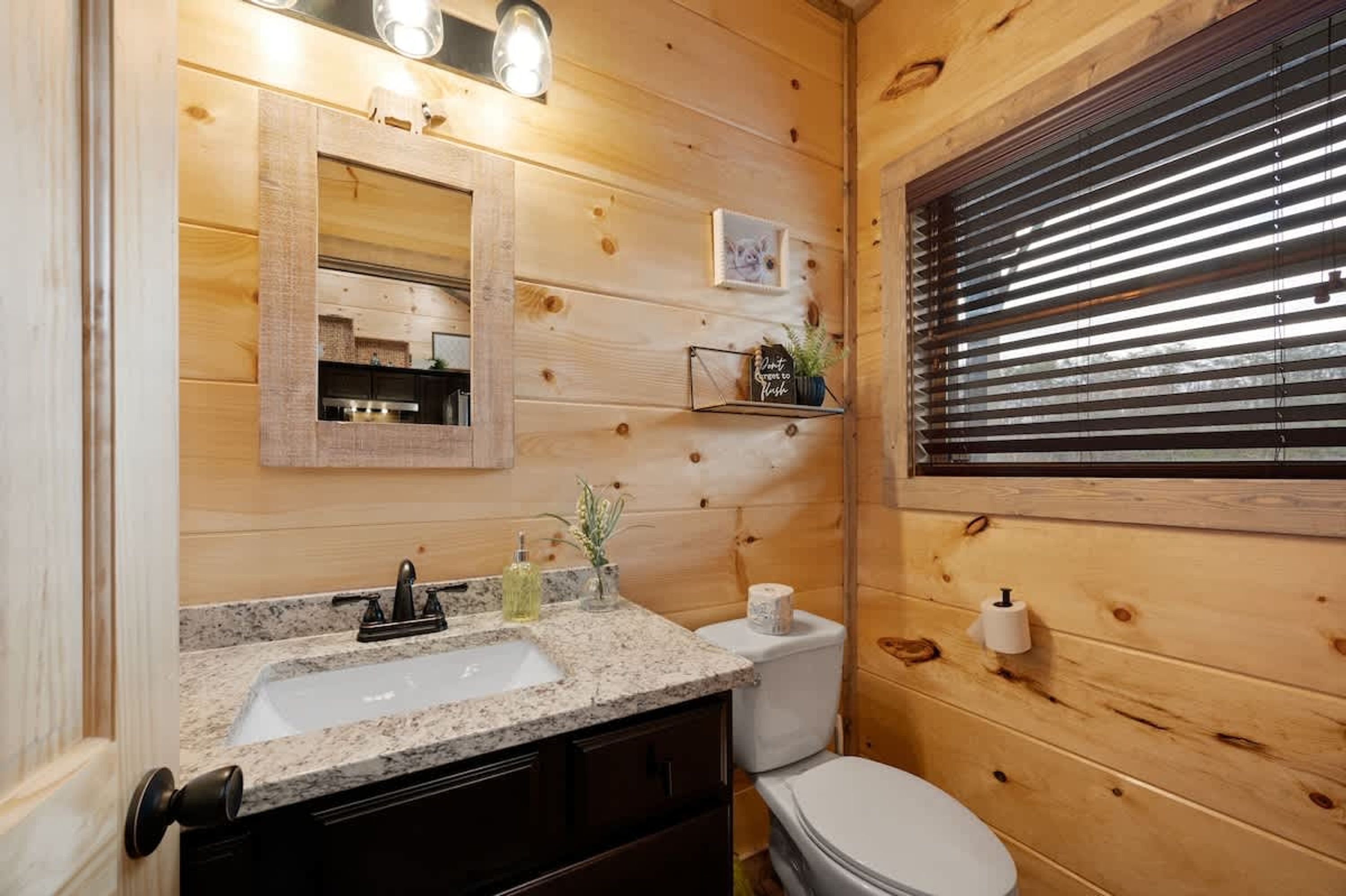 A wooden cabin bathroom with a granite sink, a mirror, a toilet, and a window with blinds.