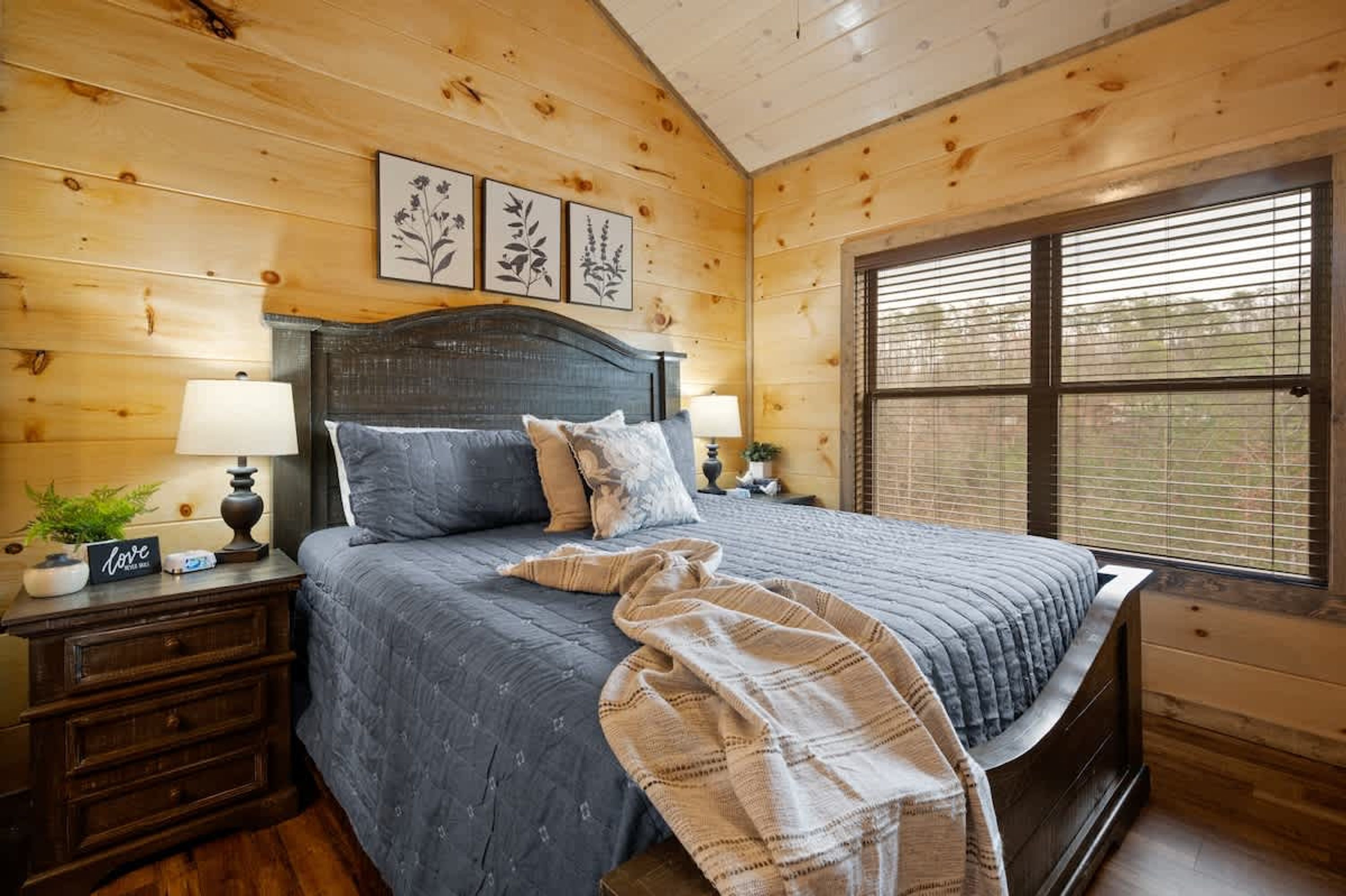 A bedroom with a wooden ceiling, a bed with a dark headboard, two bedside tables with lamps, and a window with blinds.